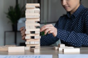 Indian businesswoman sitting at workplace desk build tower of wooden blocks play game, apply skills and creative approach close up image. Improve precision, business strategic thinking concept